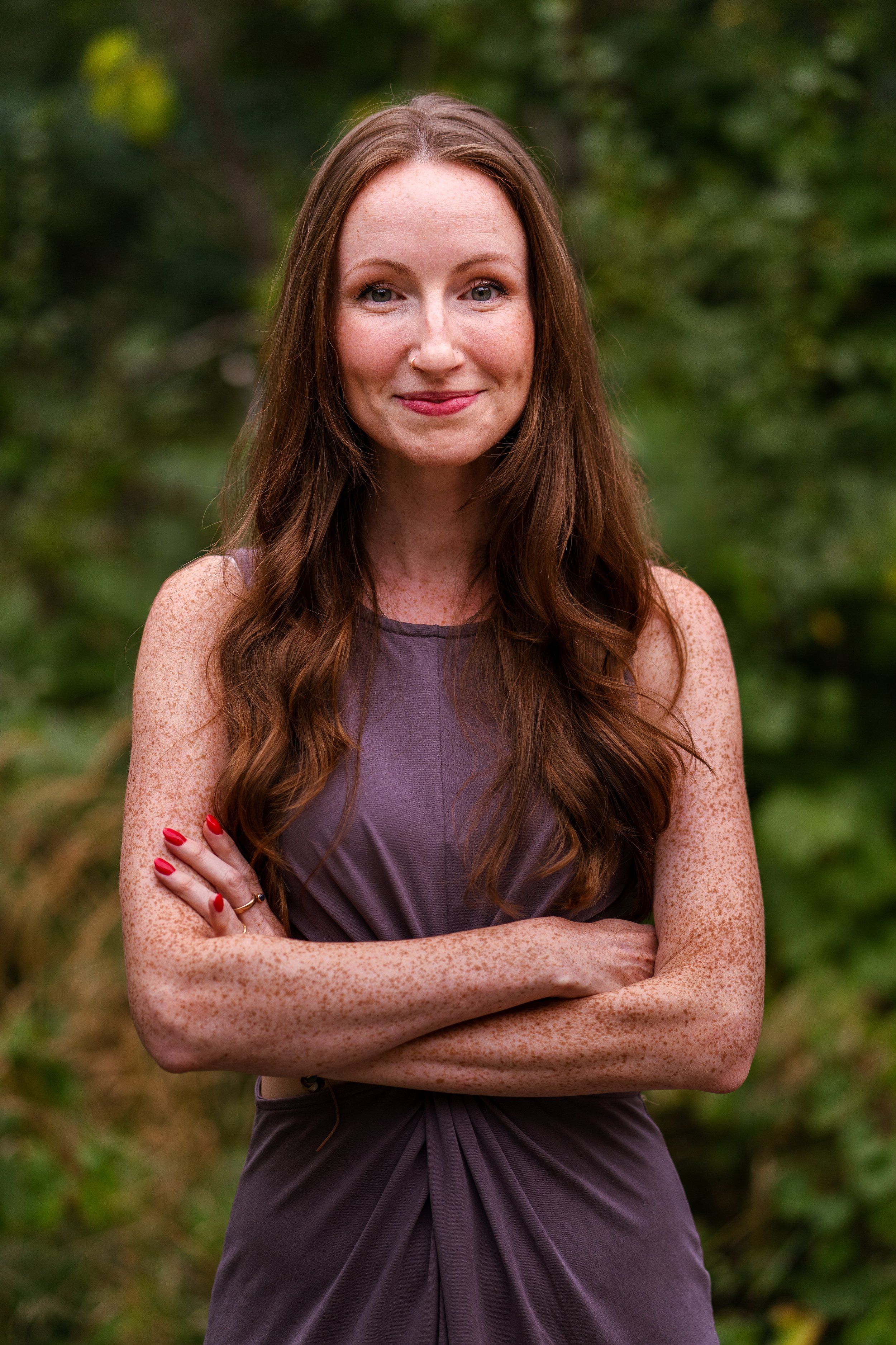 Amelia Rose, smiling, standing outdoors among green trees with her arms crossed.